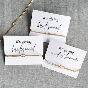 May include: Three white cards with gold bracelets, each with the phrase "it's giving" and a wedding party role: "bridesmaid" or "maid of honor". The cards are on a gray textured surface, with a gold chain in the background.