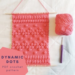 May include: A pink crochet wall hanging with a "Dynamic Dots" PDF crochet pattern title. The hanging is made with a textured stitch that creates a dotted effect. The image also shows a ball of pink yarn and a purple crochet hook.