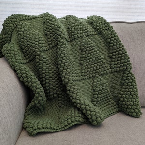 May include: A green crocheted throw blanket with a textured, bumpy pattern. The blanket is folded and draped over a couch.