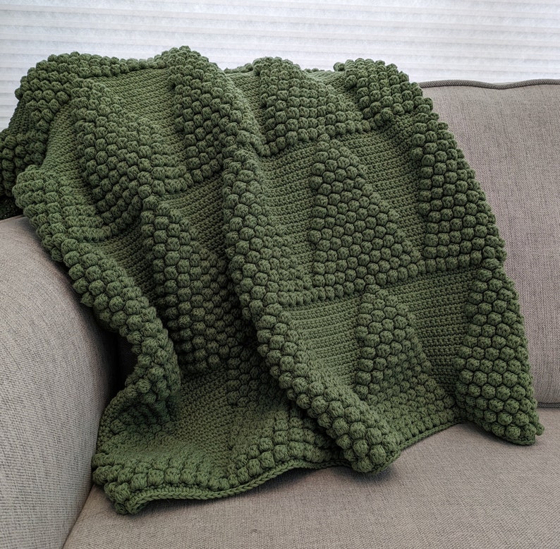May include: A green crocheted throw blanket with a textured, bumpy pattern. The blanket is folded and draped over a couch.