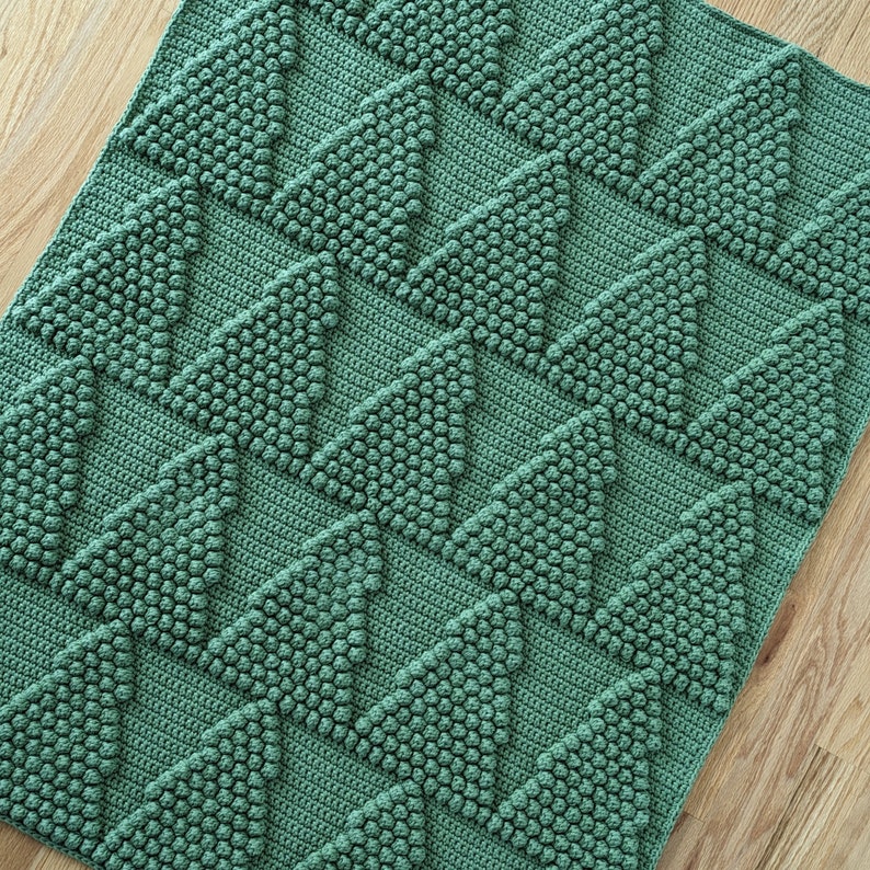 May include: A green crocheted blanket with a textured diamond pattern. The blanket is made with a repeating pattern of small, raised circles.