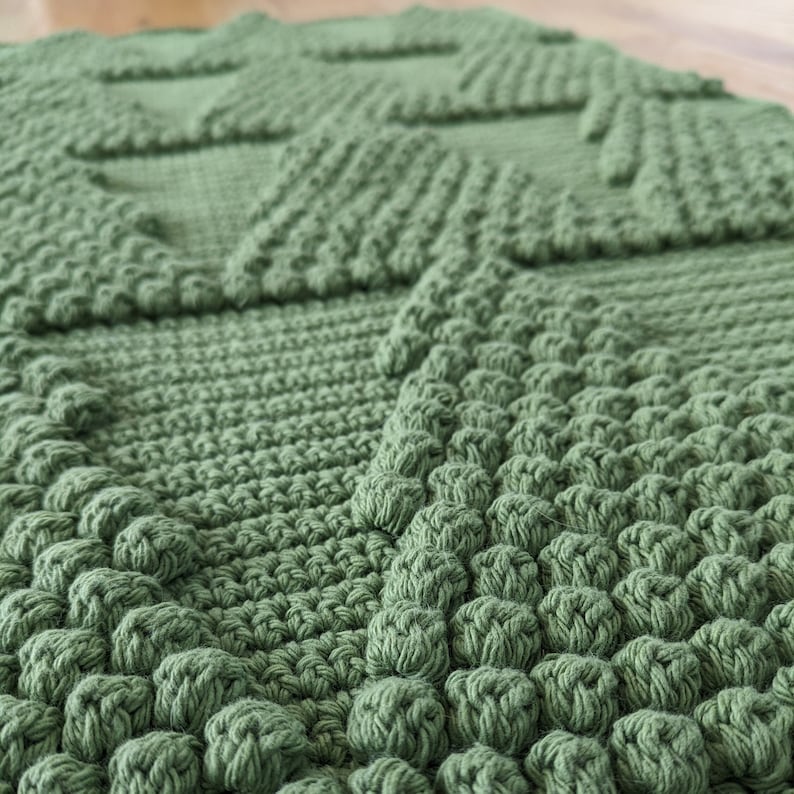 May include: A close-up of a green crocheted rug with a textured, diamond-shaped pattern. The rug is made with thick yarn and has a soft, plush feel.