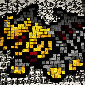 May include: A crocheted wall hanging featuring two pixelated figures, one in yellow and orange and the other in gray and black.
