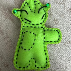Puede incluir: Un personaje hecho a mano de fieltro verde lima con detalles cosidos en negro y un contorno cosido en verde lima. El personaje tiene dos ojos verdes en forma de alfiler y está de pie. Los brazos del personaje están extendidos.