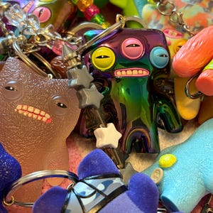 Op de afbeelding: Een verzameling kleurrijke, eigenzinnige sleutelhangers met verschillende monsterachtige karakters. De sleutelhangers hebben een verscheidenheid aan kleuren, waaronder paars, blauw, groen en geel. Sommige hebben stervormige accenten en metalen sleutelringen.