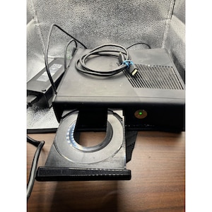 Peut inclure: Console Xbox 360 noire avec un lecteur de disque ouvert, un cordon d'alimentation et une alimentation. La console a un voyant d'alimentation vert et un câble noir connecté à l'arrière. Le lecteur de disque est argenté et a un design circulaire.