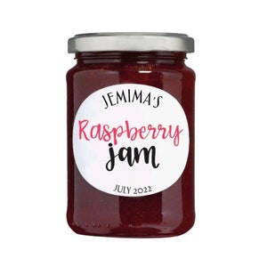 Może przedstawiać: Szklany słoik z dżemem malinowym z okrągłą białą etykietą, na której napisano "Jemima's Raspberry jam" czarnymi i różowymi literami oraz "JULY 2022" czarnymi literami.
