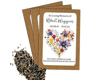 May include: A set of wildflower seed packets with a brown kraft paper backing. Each packet features a floral heart design with the text "In Loving Memory of Robert Wiggins" and dates. A pile of seeds is visible.