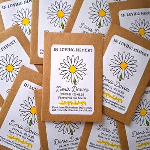 Può includere: Un mucchio di bustine di semi di carta marrone con etichette bianche. Le etichette hanno un'illustrazione di margherita in bianco e nero e il testo "In Loving Memory Doris Davies 29.09.31 - 23.01.25 Forever in our hearts Plant these Michaelmas Daisy seeds and remember Doris as they bloom."