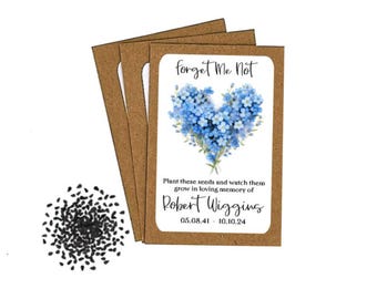 May include: Seed packets with a white label featuring a blue forget-me-not flower design in a heart shape. The text reads "forget Me Not" and "Plant these seeds and watch them grow in loving memory of Robert Wiggins 05.08.41 - 10.10.24". A pile of black seeds is also visible.