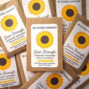 Op de afbeelding: Bruine papieren zaadzakjes met zonnebloemontwerpen en etiketten met de tekst "In Loving Memory" en "Jean Brough 09/08/1939 - 04/02/2025 Forever in our hearts Plant these seeds & remember Jean as they grow"
