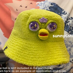 以下が含まれることがあります： ふわふわの質感の明るい黄色のクロシェの バケットハット。帽子には、紫色の目、黄色のくちばし、赤い口のクロシェの顔が付いています。帽子には「sunnyGstitches」の文字が見えます。