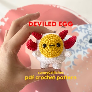 Op de afbeelding: Een gehaakt amigurumi-speelgoed in de vorm van een duivelsei. Het ei is wit met een gele dooier en rode duivelshoorns. Het speelgoed wordt in een hand gehouden. De tekst "DEVILED EGG" staat boven het speelgoed en "sunnyGstitches pdf crochet pattern" staat onder het speelgoed.