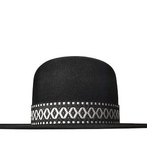 Op de afbeelding: Een zwarte hoed met een afgeronde kroon en een brede, platte rand. De basis van de kroon is voorzien van een decoratieve band met een herhalend wit geometrisch ruitpatroon op een zwarte achtergrond, rondom de gehele hoed.