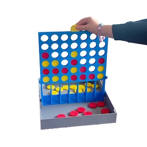May include: A blue Connect Four game with red and yellow game pieces. The game board has a grid of holes where the pieces are dropped. A hand is placing a yellow disc into the top of the board. Some red and yellow discs are in the tray.
