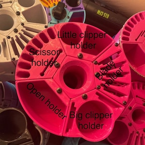 May include: A pink rotating organizer with six compartments for holding hairdressing tools. The compartments are labeled "Scissor holder", "Little clipper holder", "Big clipper holder", "Open holder", "Blade holder".