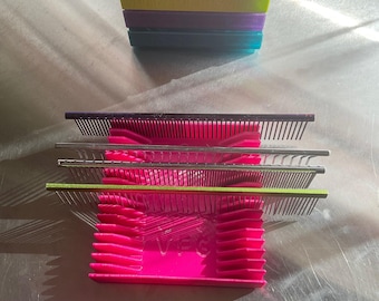 May include: A pink plastic comb holder with three silver combs stacked inside. The combs have green handles.