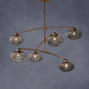 May include: A modern chandelier with six gray glass globe shades. The fixture has a brass finish and a multi-arm design. The light fixture hangs from a single point and is suitable for a dining room or living room.