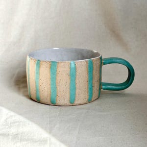 May include: A handcrafted ceramic mug featuring a speckled beige exterior with vertical turquoise stripes. The interior is light grey. The mug has a rounded turquoise handle. Ideal for tea, coffee, or other drinks.