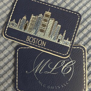 May include: Two dark blue leather coasters with gold stitching. The top coaster features a gold silhouette of the Boston skyline with the word "BOSTON" in gold lettering. The bottom coaster features a gold cursive logo "MLC" with the text "MLC GIFTS LLC" in gold lettering.