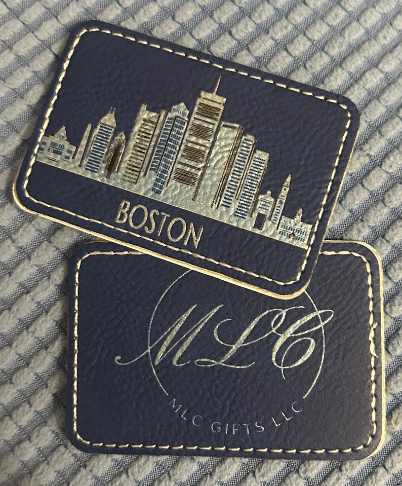 May include: Two dark blue leather coasters with gold stitching. The top coaster features a gold silhouette of the Boston skyline with the word "BOSTON" in gold lettering. The bottom coaster features a gold cursive logo "MLC" with the text "MLC GIFTS LLC" in gold lettering.