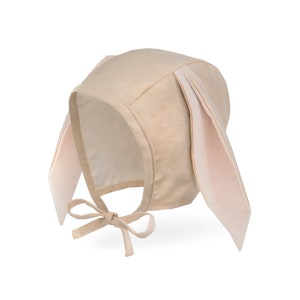 May include: A beige baby bonnet with long, floppy bunny ears. The bonnet has a rounded crown and a tie closure under the chin. The ears are a lighter shade of beige with a pink interior.