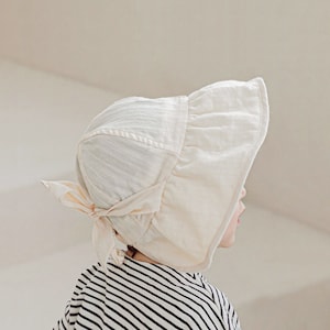 May include: A cream-colored bonnet with a ruffled brim and a bow at the back. The bonnet is made of a soft, textured fabric. The person in the image is wearing a black and white striped shirt.
