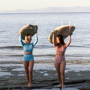 Woman in eco-friendly high-waist swim shorts with vibrant coral print, holding a surfboard at the beach. Perfect for surfing, sunbathing, and water sports, made from sustainable recycled fabric for a comfortable fit.