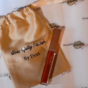 Puede incluir: Un tubo dorado de brillo de labios con el texto "Beautifully Lavish By Dori" en la etiqueta. El brillo de labios está sobre una bolsa de satén dorada.