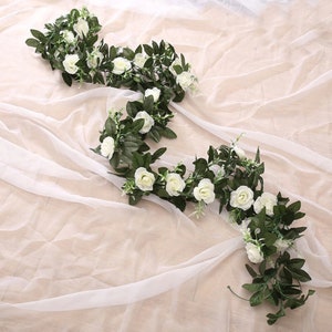Könnte beinhalten: Eine Girlande aus künstlichen weißen Rosen und grünen Blättern. Die Girlande ist über einen weißen, durchsichtigen Stoff gelegt.