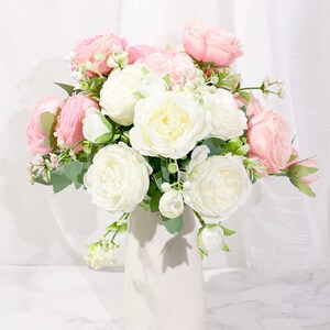 May include: A bouquet of artificial flowers in a white vase. The bouquet features a mix of white and pink roses with green leaves.