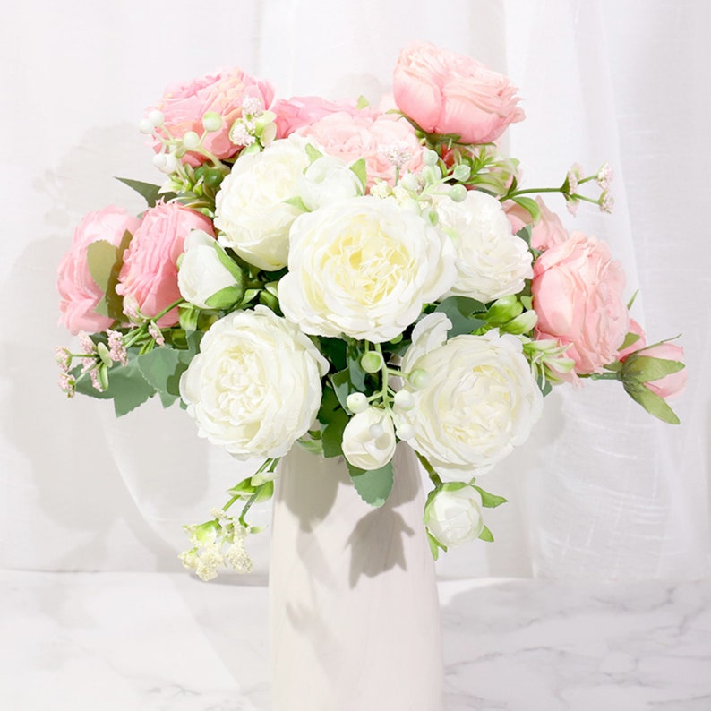 May include: A bouquet of artificial flowers in a white vase. The bouquet features a mix of white and pink roses with green leaves.