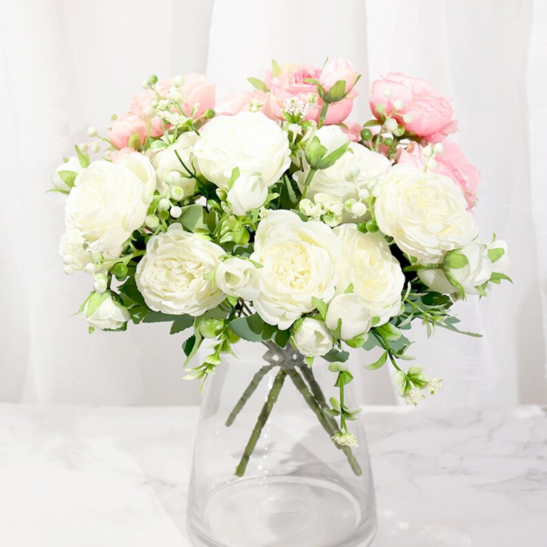 May include: A bouquet of white and pink artificial flowers in a clear glass vase. The flowers are arranged in a loose, natural style, and the vase is simple and elegant.