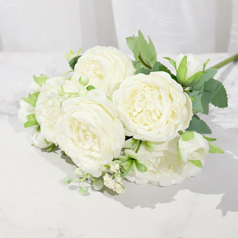 May include: A bouquet of white artificial peonies with green leaves and small white buds.