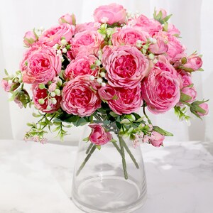 May include: A bouquet of pink artificial roses in a clear glass vase. The roses are arranged in a cluster with green leaves and small white flowers.