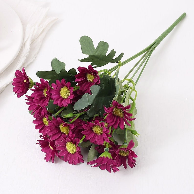 Puede incluir: Un ramo de margaritas artificiales de color p&uacute;rpura con hojas verdes. Los tallos son largos y delgados, lo que los hace ideales para usar en un jarr&oacute;n o como elemento decorativo.