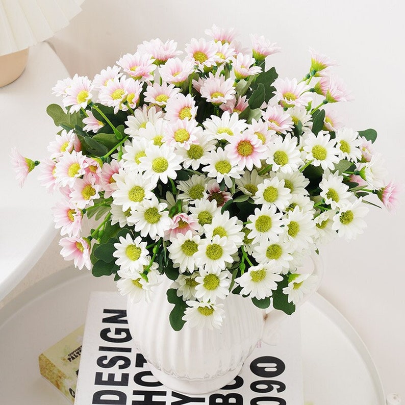 Puede incluir: Un ramo de margaritas artificiales blancas y rosas en un jarr&oacute;n de cer&aacute;mica blanco. El jarr&oacute;n tiene un dise&ntilde;o acanalado y est&aacute; sobre un libro blanco y negro.