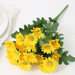 Puede incluir: Un ramo de margaritas amarillas artificiales con hojas verdes. Las flores est&aacute;n dispuestas en un racimo y tienen un aspecto realista.
