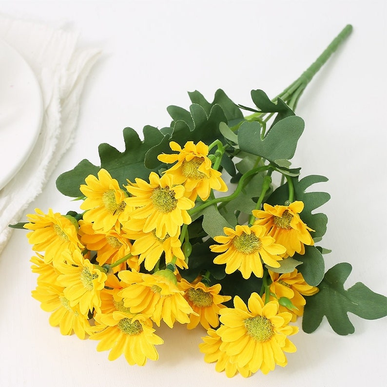 Puede incluir: Un ramo de margaritas amarillas artificiales con hojas verdes. Las flores est&aacute;n dispuestas en un racimo y tienen un aspecto realista.