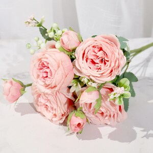 May include: A bouquet of pink artificial roses with green leaves and small white berries. The roses are arranged in a cluster, with some buds and some fully bloomed flowers.