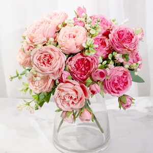 May include: A bouquet of pink and white artificial roses with green leaves and small white flowers. The roses are arranged in a clear glass vase.
