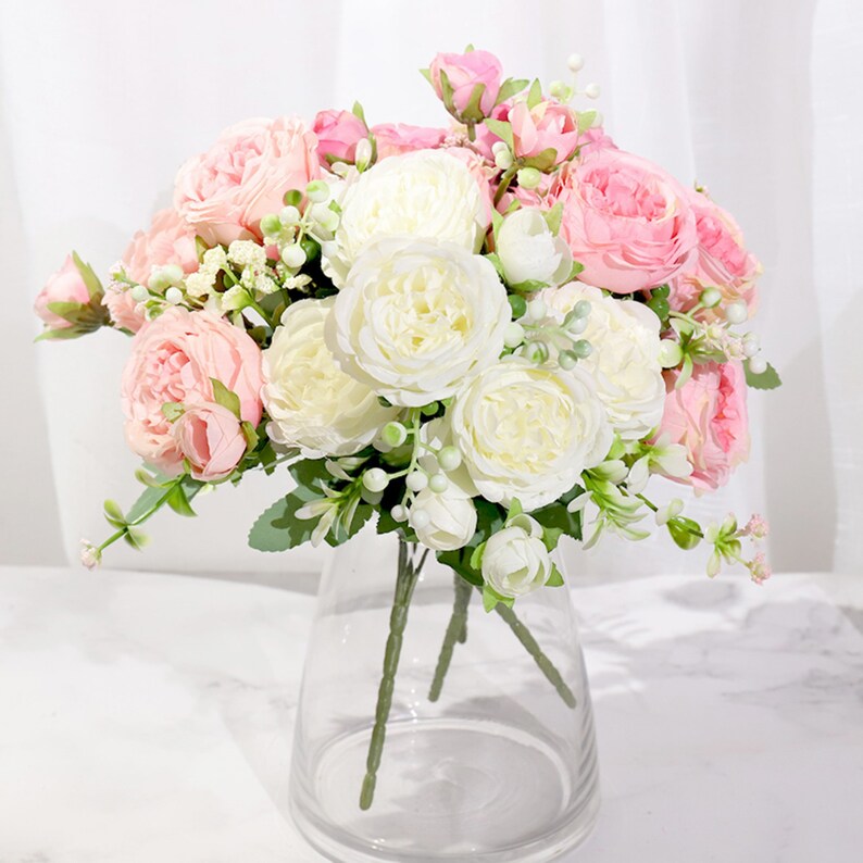 May include: A bouquet of artificial flowers in a clear glass vase. The bouquet features a mix of white and pink roses with small green leaves.