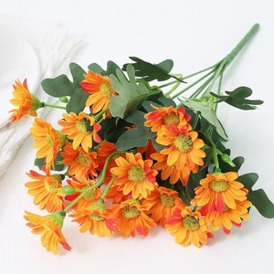 Puede incluir: Un ramo de flores de margarita artificiales de color naranja con hojas verdes. Las flores est&aacute;n dispuestas en un racimo y tienen un aspecto realista.