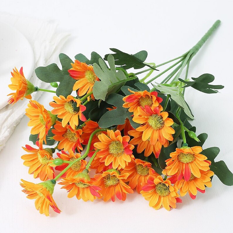 Puede incluir: Un ramo de flores de margarita artificiales de color naranja con hojas verdes. Las flores est&aacute;n dispuestas en un racimo y tienen un aspecto realista.