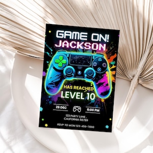 Puede incluir: Tarjeta de invitación negra con un gráfico de mando de videojuegos azul y verde neón. El texto dice "GAME ON! JACKSON HAS REACHED LEVEL 10" con detalles del evento. El diseño incluye salpicaduras de pintura coloridas.