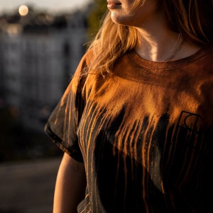 Könnte beinhalten: Eine Frau mit Sonnenbrille und einem schwarzen und orangefarbenen Batik-T-Shirt. Das Shirt hat ein vertikales Tropfmuster mit schwarzer und orangefarbener Farbe.