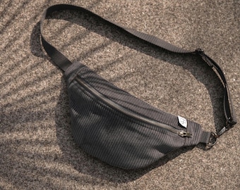 May include: A dark gray fanny pack featuring a ribbed texture, a horizontal zippered closure, and an adjustable black nylon strap. A small white rectangular fabric tag with black text is attached to the side of the zipper opening.