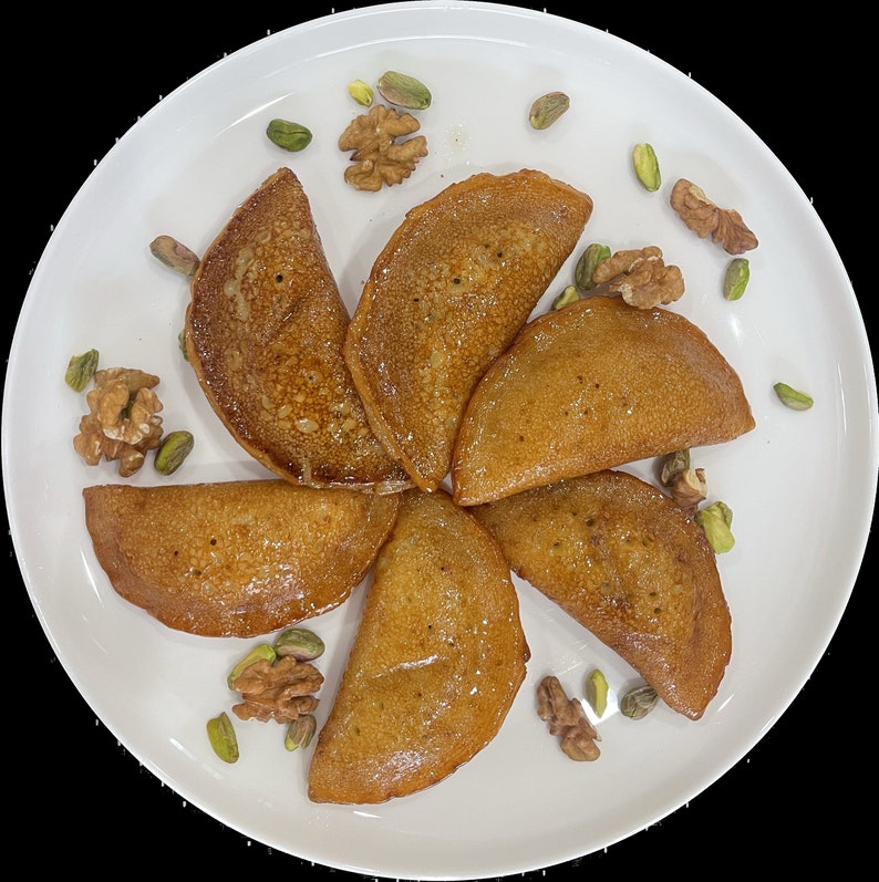 May include: A white plate with seven golden brown crescent-shaped pastries topped with a drizzle of honey. The plate is decorated with pistachios and walnuts.
