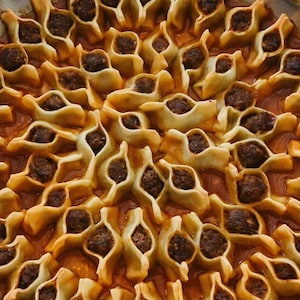 May include: Close-up of a dish of Turkish manti, small dumplings filled with seasoned ground meat. The dumplings are arranged in a pattern and sit in a tomato-based sauce. The manti are golden brown and the filling is dark brown.