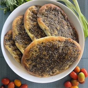 May include: A white bowl filled with five flatbreads topped with a mixture of sesame seeds and zaatar. The flatbreads are arranged in a circle, with the largest one in the center. The bowl is sitting on a blue and white patterned surface. There are green onions and red tomatoes scattered around the bowl.